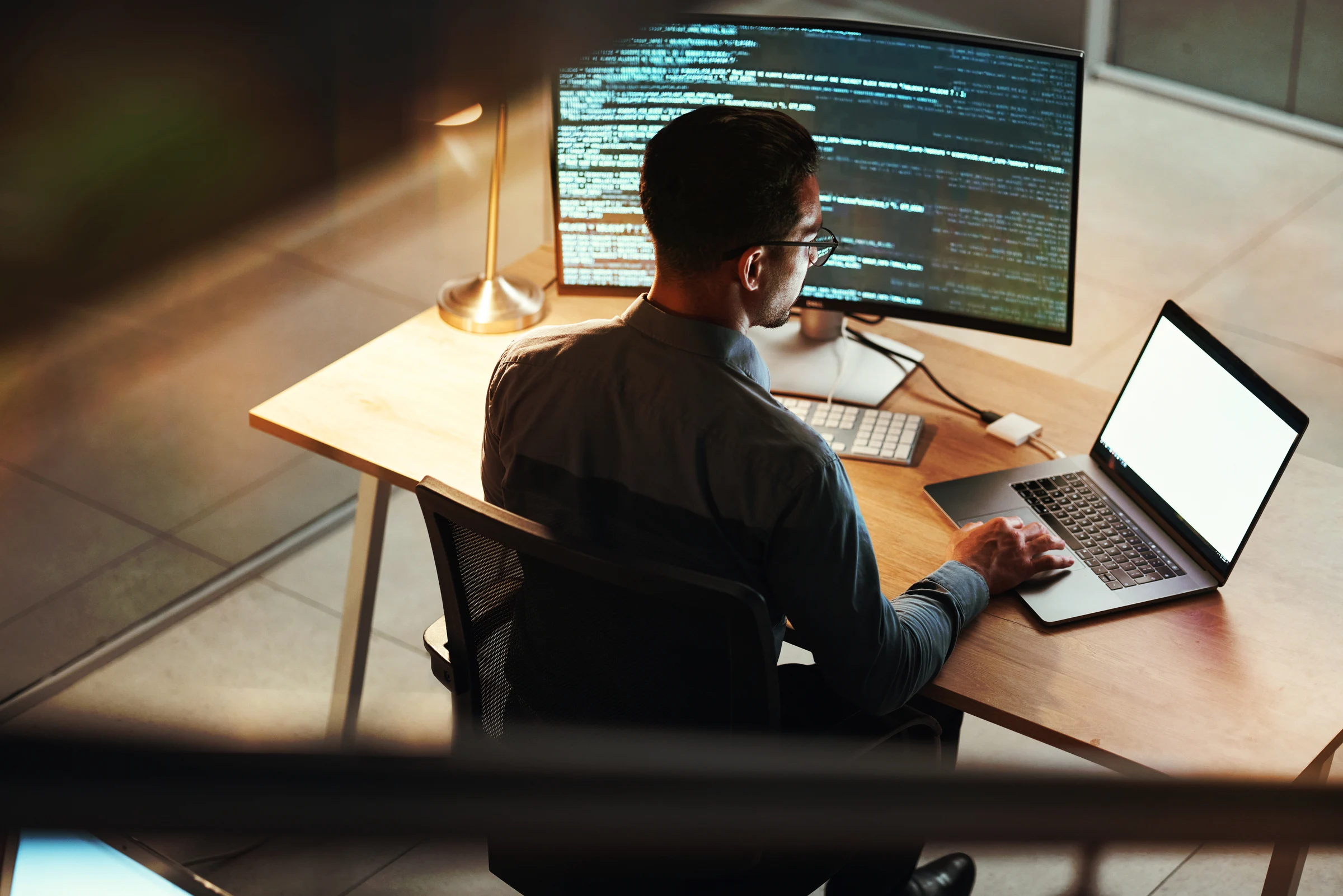 An office worker sits at a desk with their back to the camera, looking at a laptop. A larger desktop screen is in front of them.