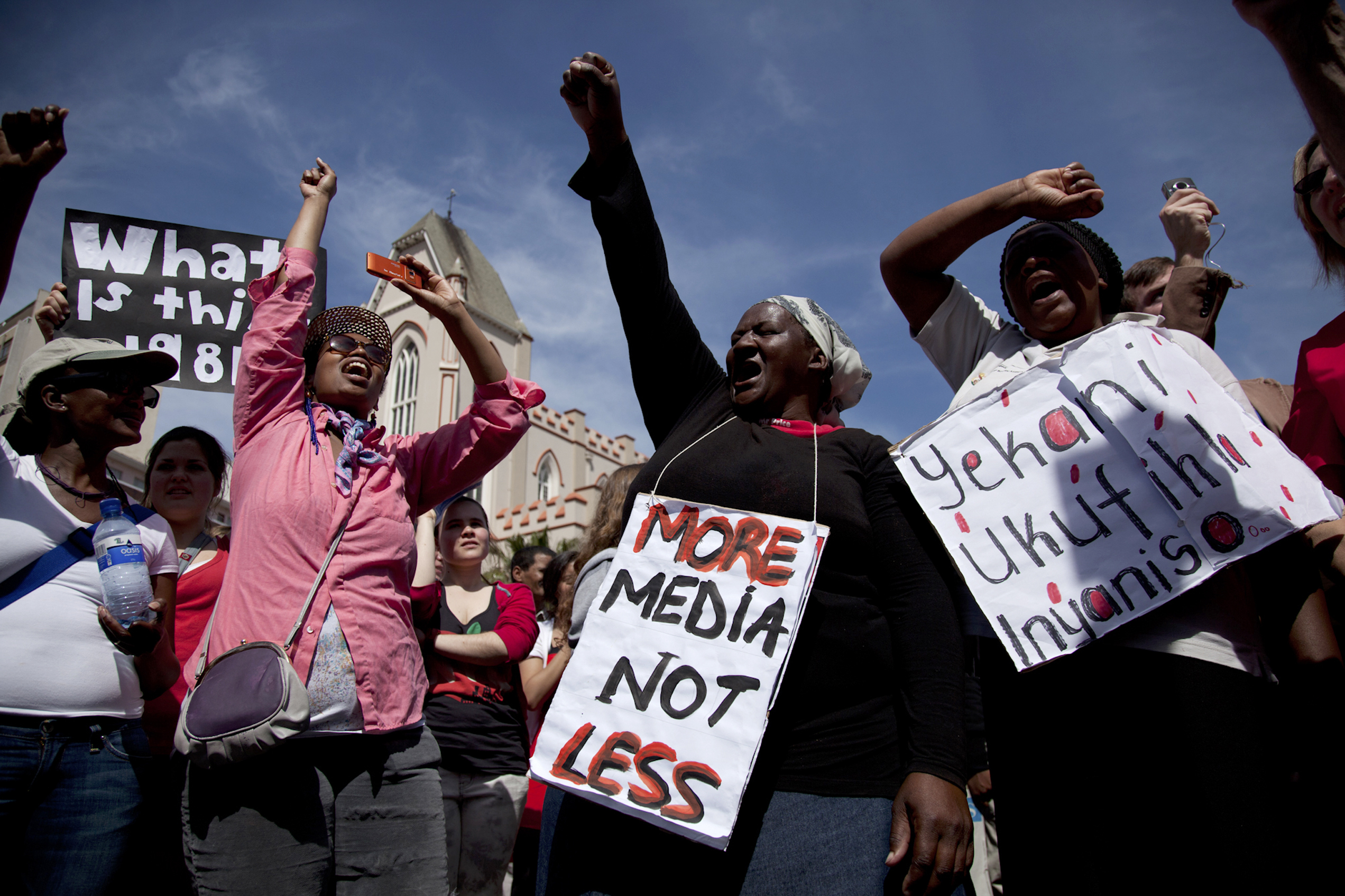 Protesters raise their fists outside a church, holding signs reading "More media, not less" against a blue sky.