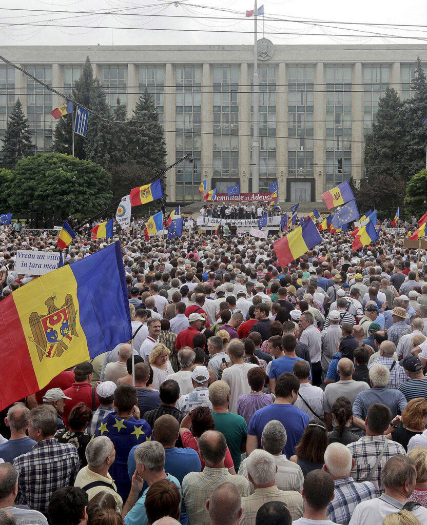 A large crowd standing outside a state building, several of them holding large Moldovan flags above their heads.